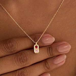 May include: A gold necklace with a rectangular pendant, set with a red and a pink gemstone. The pendant hangs from a fine gold chain. The necklace is shown against a neutral backdrop.