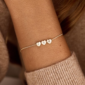 Op de afbeelding: Een fijne goudkleurige kettingarmband met drie kleine, hartvormige bedels op een rij. Elk hartje is gegraveerd met een enkele hoofdletter, namelijk E, N en K, die in het midden op de voorzijde van het metaal zijn geplaatst.