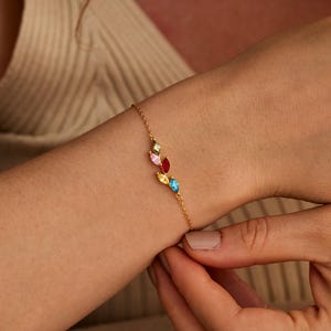 Könnte beinhalten: Ein goldfarbenes Armband mit bunten Edelsteinen. Die Edelsteine in Rosa, Gelb, Blau und Grün sind in einem blattähnlichen Muster angeordnet. Das Armband wird an einem Handgelenk gezeigt, wodurch sein elegantes Design hervorgehoben wird.