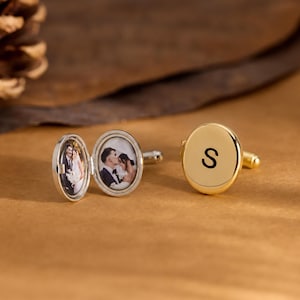 May include: A pair of gold-toned oval cufflinks. One cufflink is closed and features the black letter S engraved on its surface. The other cufflink is open, revealing a small oval photograph of a couple inside the locket-style frame.