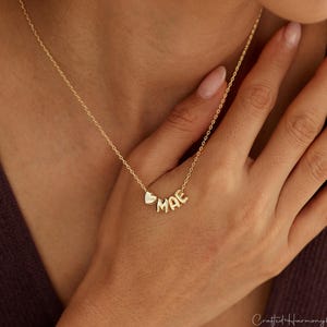 May include: A gold-toned necklace featuring a delicate chain and a pendant consisting of a small heart charm followed by the letters M, A, and E in a rounded, metallic font. The necklace rests against the neck, showcasing the gold-coloured metal finish.