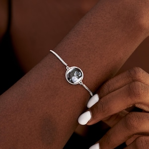 May include: A silver-toned adjustable chain bracelet featuring a circular pendant. The pendant contains a small, black and white circular image of an ultrasound scan. The bracelet has a delicate link design and a secure, sliding clasp mechanism.