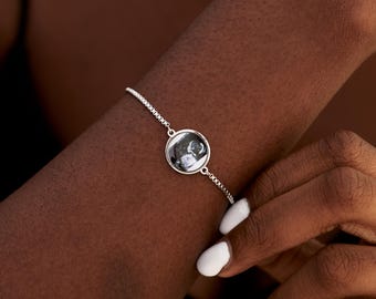 May include: A silver-toned adjustable chain bracelet featuring a circular pendant. The pendant contains a small, black and white circular image of an ultrasound scan. The bracelet has a delicate link design and a secure, sliding clasp mechanism.
