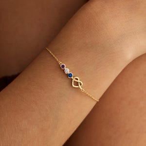 May include: A delicate gold bracelet featuring three gemstones in purple, white, and blue, and a gold infinity heart charm. The bracelet is displayed on a person's arm.
