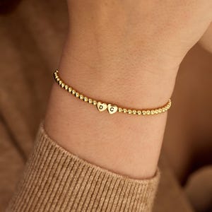 Op de afbeelding: Een armband bestaande uit kleine, uniforme en gepolijste goudkleurige kralen. Het sieraad is voorzien van twee hartvormige bedels, elk gegraveerd met een enkele letter, namelijk een D en een C. De kralen vormen een aaneengesloten band rond de pols.