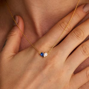 Op de afbeelding: Een gouden ketting met een hanger bestaande uit twee peervormige stenen die aan elkaar zijn bevestigd. De ene steen is diepblauw van kleur en de andere is een heldere, kleurloze steen. De hanger is gecentreerd aan een fijne, dunne gouden schakelketting.