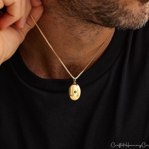 Pode incluir: Colar com pingente oval dourado com uma pequena pedra verde. O pingente pende de uma delicada corrente dourada. A pessoa está vestindo uma camiseta preta.