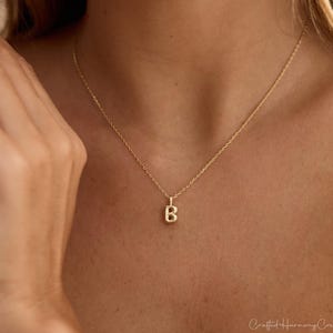 May include: A gold-toned necklace featuring a delicate chain and a small, capital letter B pendant. The pendant has a textured, slightly rounded surface and hangs centrally from the thin, polished metal chain resting against the skin.