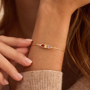 May include: A delicate gold chain bracelet featuring a central pea pod charm. The charm contains three small, round, faceted gemstones in vibrant shades of red, yellow, and purple, set horizontally within a polished gold-toned metal frame.