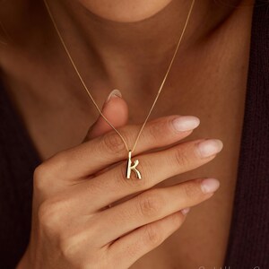 Könnte beinhalten: Eine goldfarbene Halskette mit einer zarten Kette und einem kleinen, polierten Anhänger in Form des Buchstabens K. Der Anhänger hat eine glatte, abgerundete Oberfläche und hängt mittig an der dünnen Metallkette am Schlüsselbein.