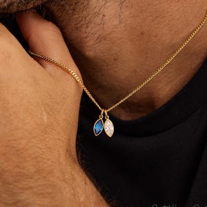 May include: A gold-colored chain necklace with two leaf-shaped pendants. One pendant features a blue gemstone, and the other has a clear gemstone. The necklace is worn around the neck, with the pendants resting on the chest.