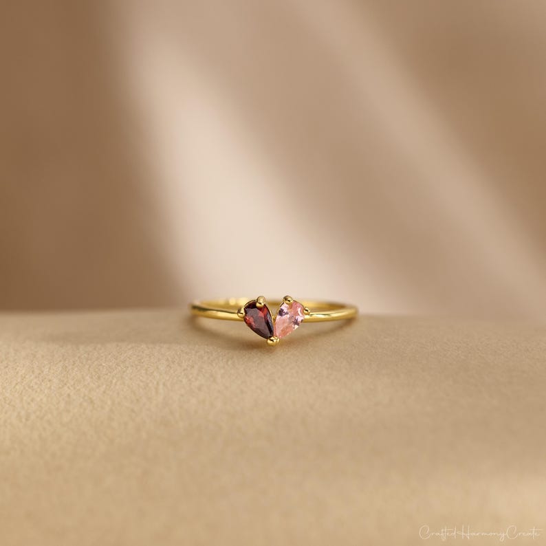 K&ouml;nnte beinhalten: Ein goldener Ring mit zwei birnenf&ouml;rmigen Edelsteinen, die zusammen eine Herzform bilden. Ein Edelstein ist ein tiefroter Granat, der andere ein hellrosa Stein. Das Band ist d&uuml;nn und aus poliertem Gold, was ein zartes und feines Aussehen ergibt.