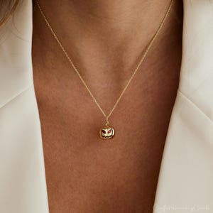 May include: A gold-colored necklace with a pumpkin pendant. The pendant has a carved face with red accents for the eyes. The necklace is worn against a white blazer.