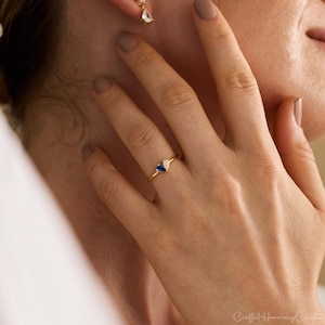 K&ouml;nnte beinhalten: Ein goldener Ring mit zwei kleinen tropfenf&ouml;rmigen Steinen, einem tiefblauen und einem klaren wei&szlig;en, die nebeneinander auf einem d&uuml;nnen Ringband sitzen. Der Ring wird an einem Finger getragen und ein passender Ohrring ist am Ohrl&auml;ppchen zu sehen.