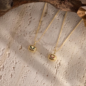 May include: Two gold-colored necklaces with pendant charms. One pendant features green gemstones, the other red. The pendants are pumpkin-shaped with carved details. The necklaces are displayed on a textured, light-colored surface.