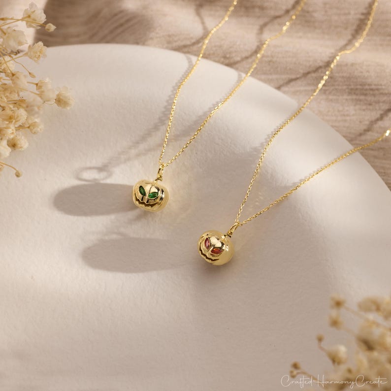 May include: Two gold-colored necklaces with pumpkin-shaped pendants. One pendant has green and red accents, while the other has red and orange accents. The necklaces are on a white surface with dried flowers in the background.