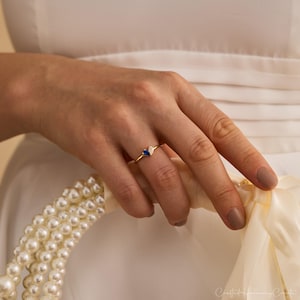 K&ouml;nnte beinhalten: Ein Goldring mit zwei kleinen birnenf&ouml;rmigen Steinen, einem tiefblauen und einem klaren wei&szlig;en, die nebeneinander auf einem d&uuml;nnen Goldband sitzen. Der Ring wird an einem Finger getragen, &uuml;ber einer wei&szlig;en Stoffoberfl&auml;che mit wei&szlig;en Perlen.