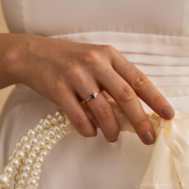 K&ouml;nnte beinhalten: Ein Goldring mit zwei kleinen birnenf&ouml;rmigen Steinen, einem tiefblauen und einem klaren wei&szlig;en, die nebeneinander auf einem d&uuml;nnen Goldband sitzen. Der Ring wird an einem Finger getragen, &uuml;ber einer wei&szlig;en Stoffoberfl&auml;che mit wei&szlig;en Perlen.