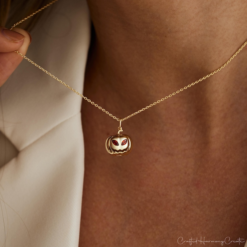 May include: A gold-colored necklace featuring a pumpkin pendant with red accents for the eyes. The pendant is detailed with carved features and hangs from a delicate chain. The necklace is shown against a neutral background.