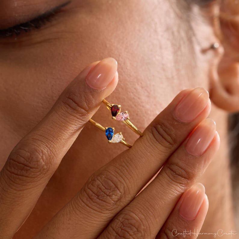 K&ouml;nnte beinhalten: Zwei goldfarbene Ringe, die jeweils mit zwei tropfenf&ouml;rmigen Edelsteinen besetzt sind. Der obere Ring hat einen dunkelroten und einen hellrosa Stein. Der untere Ring zeigt einen tiefblauen und einen klaren wei&szlig;en Stein auf d&uuml;nnen Ringen.