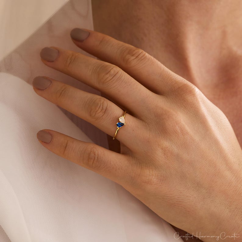 K&ouml;nnte beinhalten: Ein d&uuml;nner Goldring mit zwei kleinen, nebeneinander gesetzten birnenf&ouml;rmigen Steinen. Ein Stein hat eine tiefe Saphirblaue Farbe, der andere ist ein klarer, farbloser Stein. Die Ringschiene besteht aus einem einfachen, polierten Golddraht.