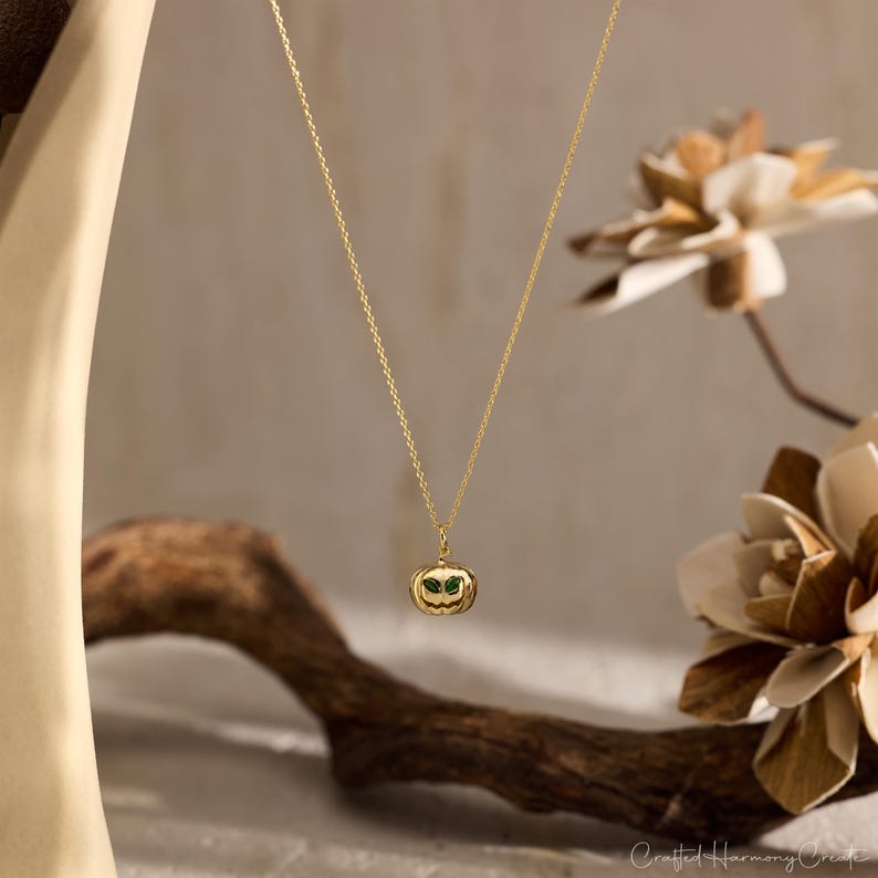 May include: A gold-colored necklace with a pumpkin pendant. The pendant has green accents for the eyes and is suspended from a delicate chain. The background features a wooden branch and decorative flowers.