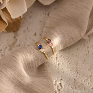 K&ouml;nnte beinhalten: Zwei Goldringe, die jeweils mit zwei birnenf&ouml;rmigen Edelsteinen besetzt sind. Ein Ring zeigt einen blauen und einen klaren Stein, w&auml;hrend der andere Ring einen dunkelvioletten und einen hellrosa Stein aufweist. Die Ringe haben d&uuml;nne Goldb&auml;nder.