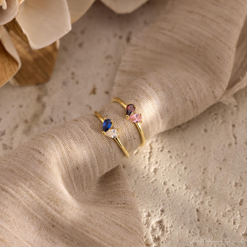 K&ouml;nnte beinhalten: Zwei Goldringe, die jeweils mit zwei birnenf&ouml;rmigen Edelsteinen besetzt sind. Ein Ring zeigt einen blauen und einen klaren Stein, w&auml;hrend der andere Ring einen dunkelvioletten und einen hellrosa Stein aufweist. Die Ringe haben d&uuml;nne Goldb&auml;nder.