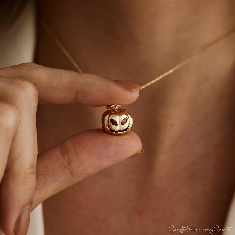 May include: A gold-colored pumpkin pendant necklace with a delicate chain. The pumpkin has carved-out eyes and a mouth, with red accents in the eye sockets. The necklace is held between fingers against a blurred background.