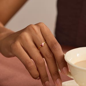 K&ouml;nnte beinhalten: Ein goldener Ring mit zwei kleinen, birnenf&ouml;rmigen Edelsteinen, die nebeneinander auf einem d&uuml;nnen Ringband sitzen. Ein Edelstein ist tiefrot, der andere zartrosa. Der Ring wird an einem Finger getragen und befindet sich neben einer wei&szlig;en Tasse.
