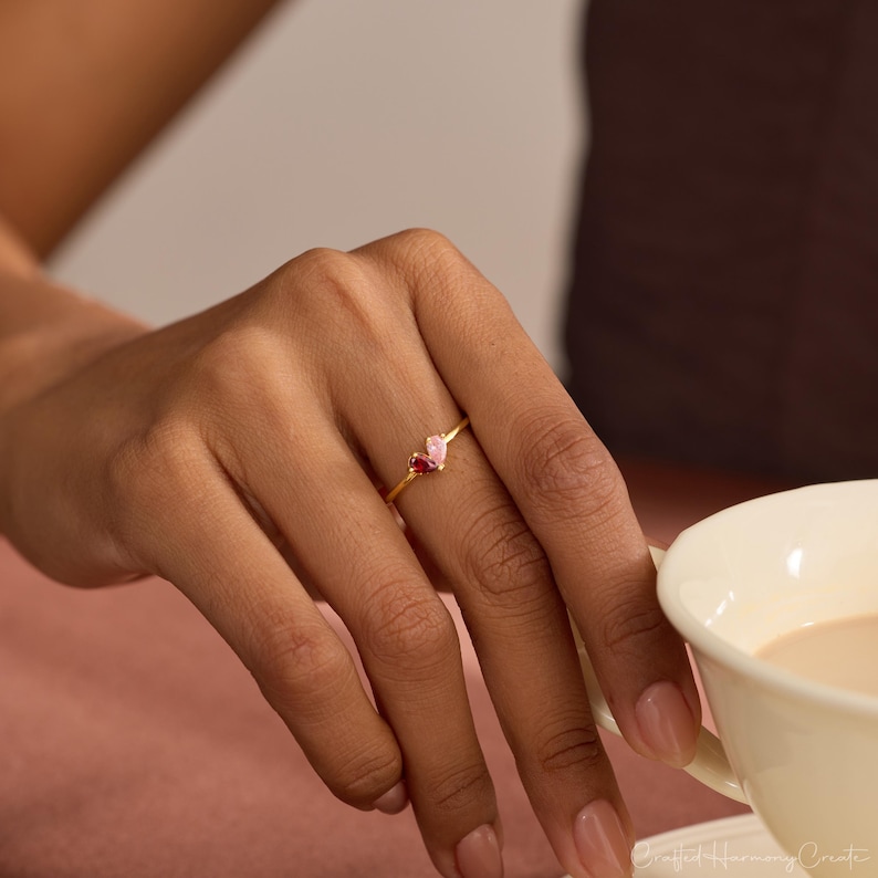 K&ouml;nnte beinhalten: Ein goldener Ring mit zwei kleinen, birnenf&ouml;rmigen Edelsteinen, die nebeneinander auf einem d&uuml;nnen Ringband sitzen. Ein Edelstein ist tiefrot, der andere zartrosa. Der Ring wird an einem Finger getragen und befindet sich neben einer wei&szlig;en Tasse.