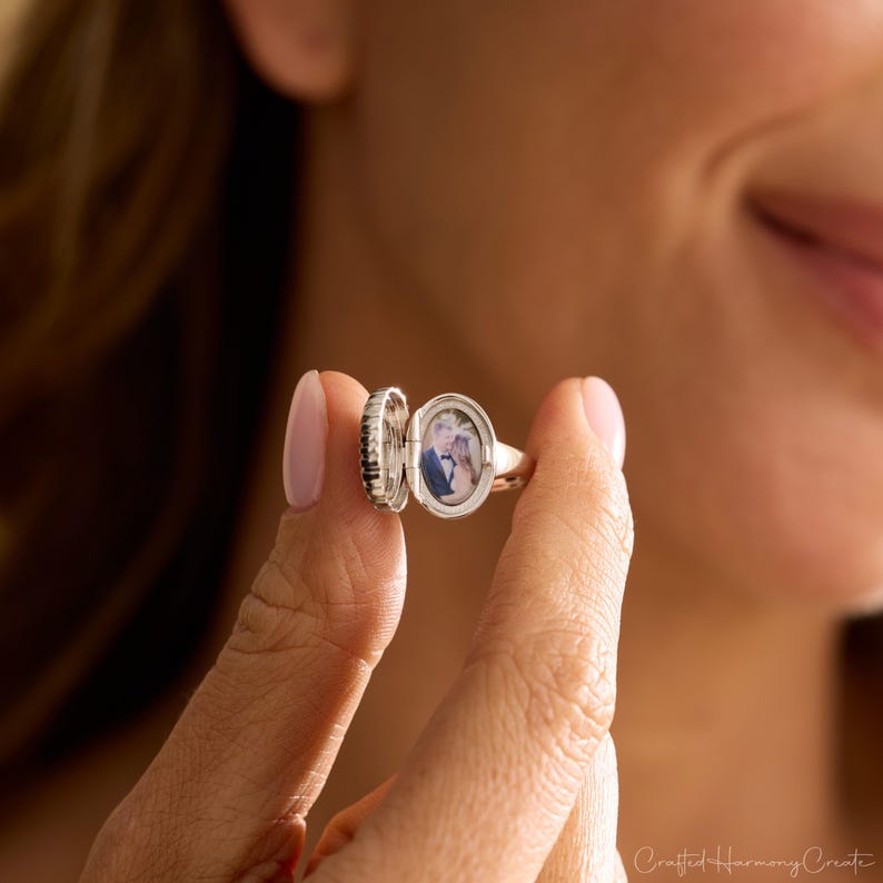 May include: A silver locket ring with an oval-shaped compartment, open to reveal a small photograph. The ring is held between fingers, showcasing its design. The ring's band is smooth, while the locket has a textured edge.