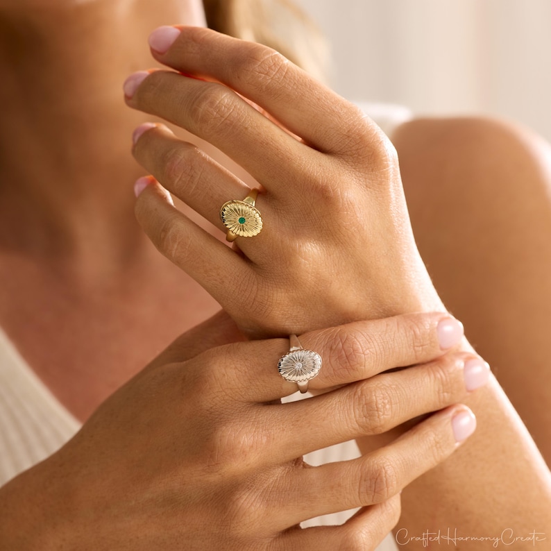 May include: Two gold-colored rings with oval-shaped faces. One ring features a small green stone in the center, while the other has a textured design. The rings are worn on fingers.