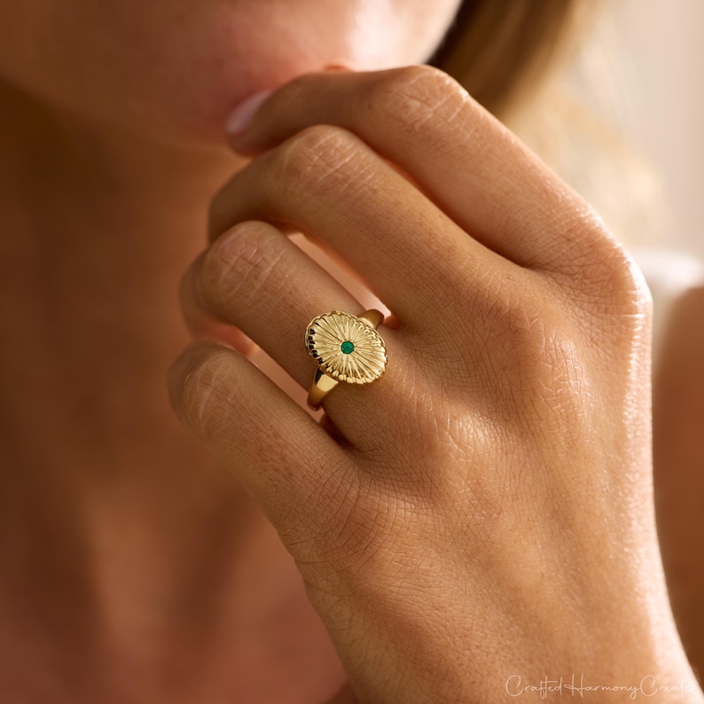 May include: A gold ring with an oval-shaped face featuring a sunburst design and a small, round, green gemstone in the center. The ring is on a finger, showcasing its intricate details and elegant style.