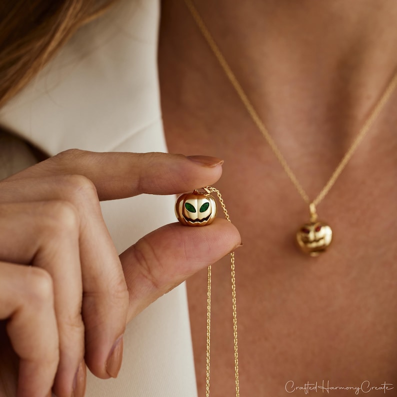 May include: A gold-colored pumpkin pendant necklace with green and black accents. The necklace is held up by a person's fingers. Another similar necklace is visible in the background.