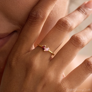 K&ouml;nnte beinhalten: Ein goldfarbener Ring mit zwei tropfenf&ouml;rmigen Edelsteinen, die nebeneinander angeordnet sind. Ein Edelstein ist tiefrot, der andere blassrosa. Der Ring hat eine d&uuml;nne, glatte Ringschiene, die den Finger umschlie&szlig;t und ein zartes Design zeigt.
