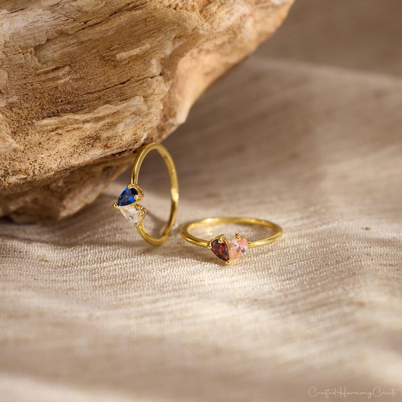 K&ouml;nnte beinhalten: Zwei goldene Ringe mit tropfenf&ouml;rmigen Edelsteinen. Ein Ring zeigt einen blauen und einen klaren Stein, w&auml;hrend der andere Ring einen dunkelroten und einen hellrosa Stein aufweist. Beide Ringe haben d&uuml;nne, polierte Goldb&auml;nder und Krappenfassungen.