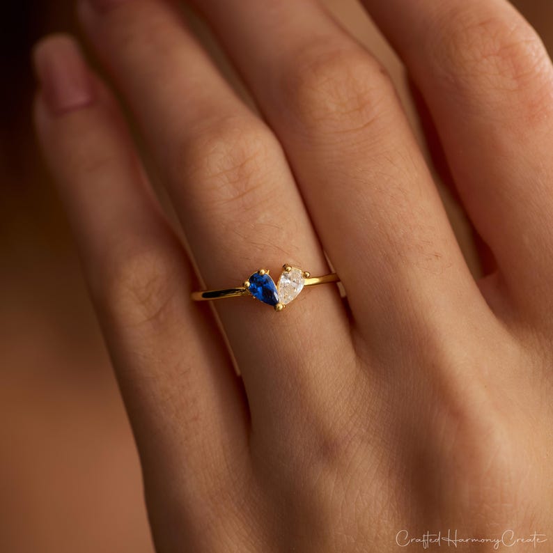 K&ouml;nnte beinhalten: Ein goldener Ring mit zwei birnenf&ouml;rmigen Steinen, die zusammen eine Herzform bilden. Ein Stein ist tiefblau saphirfarben, der andere ist ein klarer wei&szlig;er Stein. Das Ringband besteht aus d&uuml;nnem, poliertem Goldmetall und wirkt zart.