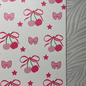May include: A white rectangular surface featuring a repeating pattern of pink cherries tied with pink bows, interspersed with small pink stars and additional pink bows. The design uses shades of light and dark pink on a clean white background.