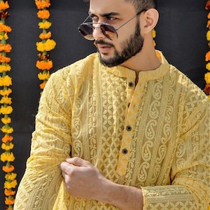 May include: A yellow embroidered caftan with a button closure. The caftan has a paisley pattern and is made of a sheer fabric. The model is wearing sunglasses and has a beard.