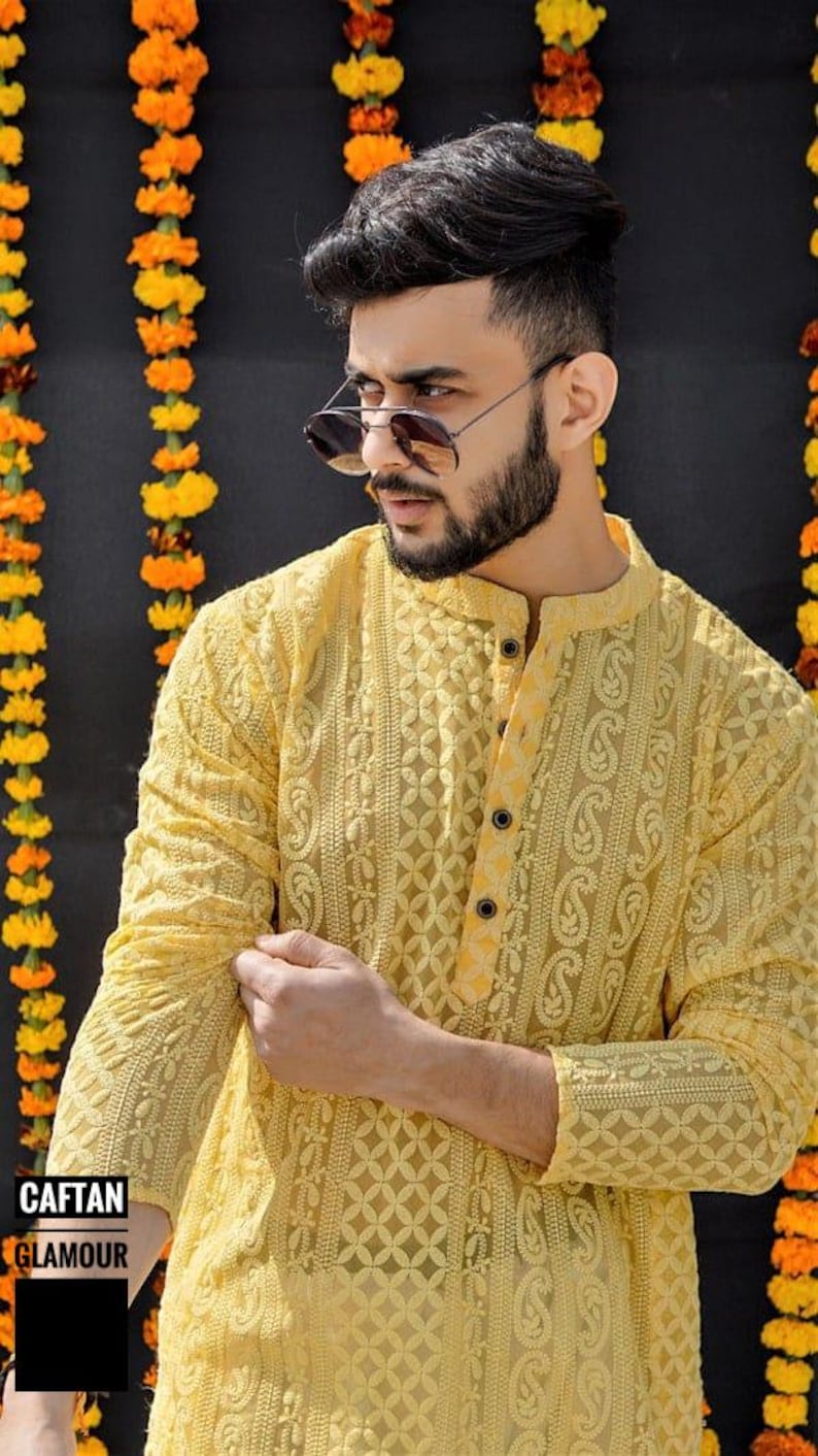 May include: A yellow embroidered caftan with a button closure. The caftan has a paisley pattern and is made of a sheer fabric. The model is wearing sunglasses and has a beard.