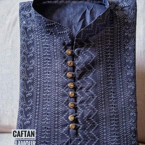 May include: A navy blue embroidered tunic with a mandarin collar and gold-colored buttons. The tunic features intricate white stitching details and the text "CAFTAN GLAMOUR" in a white font.