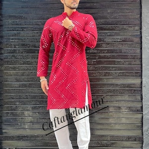 Puede incluir: Una kurta roja de manga larga con lentejuelas blancas y una abertura lateral blanca. La kurta se lleva con pantalones blancos y zapatos marrones. El texto "Caftanglamour" es visible en la imagen.