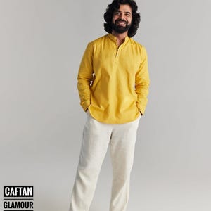May include: A man wearing a yellow long-sleeved shirt with a mandarin collar and white pants. He is smiling and has his hands in his pockets.