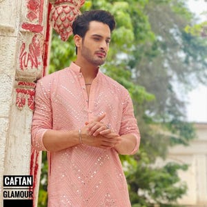 May include: A man wearing a pink long-sleeved shirt with a white embroidered design and silver embellishments. The shirt has a mandarin collar and a button closure. The man is standing in front of a red and white wall.