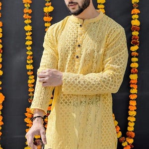 May include: A yellow long-sleeved shirt with intricate embroidery and a button-up collar. The shirt is worn by a person standing in front of a wall decorated with yellow flower garlands.