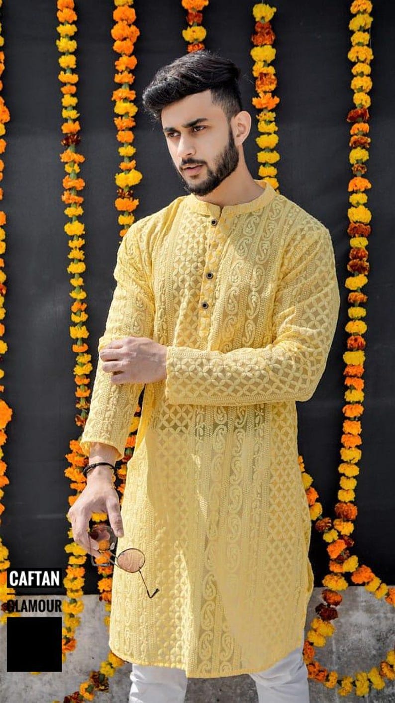 May include: A yellow long-sleeved shirt with intricate embroidery and a button-up collar. The shirt is worn by a person standing in front of a wall decorated with yellow flower garlands.