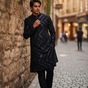 May include: A black long-sleeved tunic with a geometric pattern of silver embellishments. The tunic is paired with black pants and shoes. The outfit is worn by a person standing on a cobblestone street.