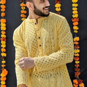 May include: A man wearing a yellow embroidered kurta with a button-up closure. He is standing in front of a wall decorated with yellow flower garlands.