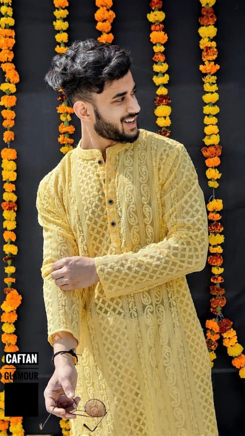 May include: A man wearing a yellow embroidered kurta with a button-up closure. He is standing in front of a wall decorated with yellow flower garlands.